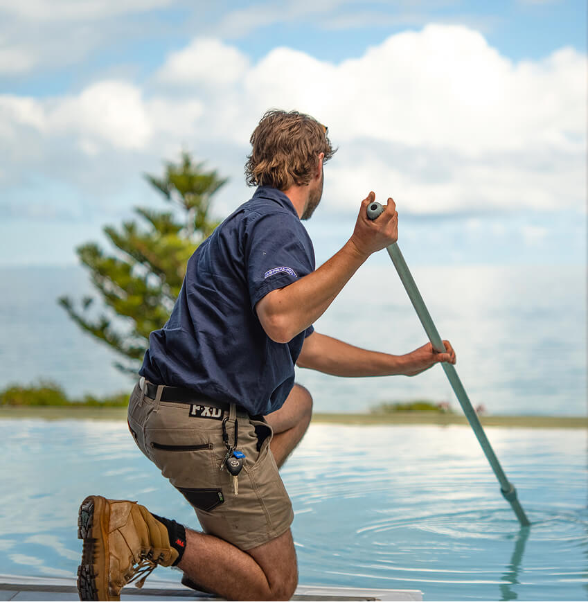 Technician kneeling on the edge of an infinity pool, cleaning it with a long-handled pole
