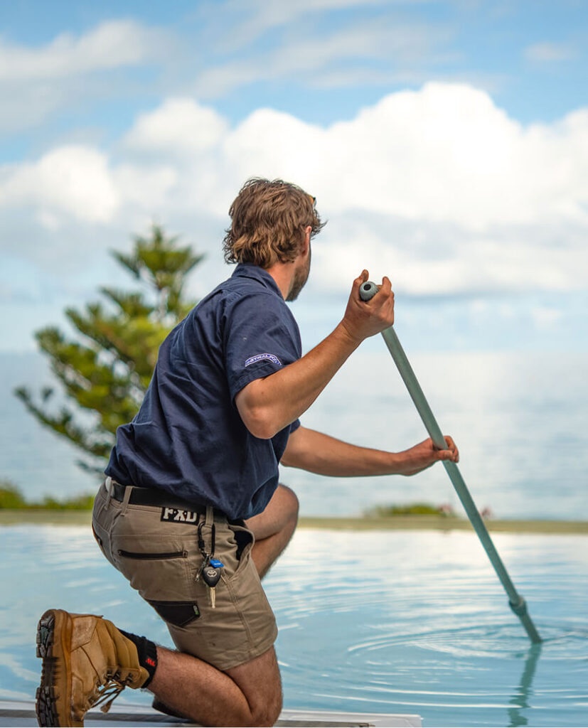 Technician kneeling on the edge of an infinity pool, cleaning it with a long-handled pole